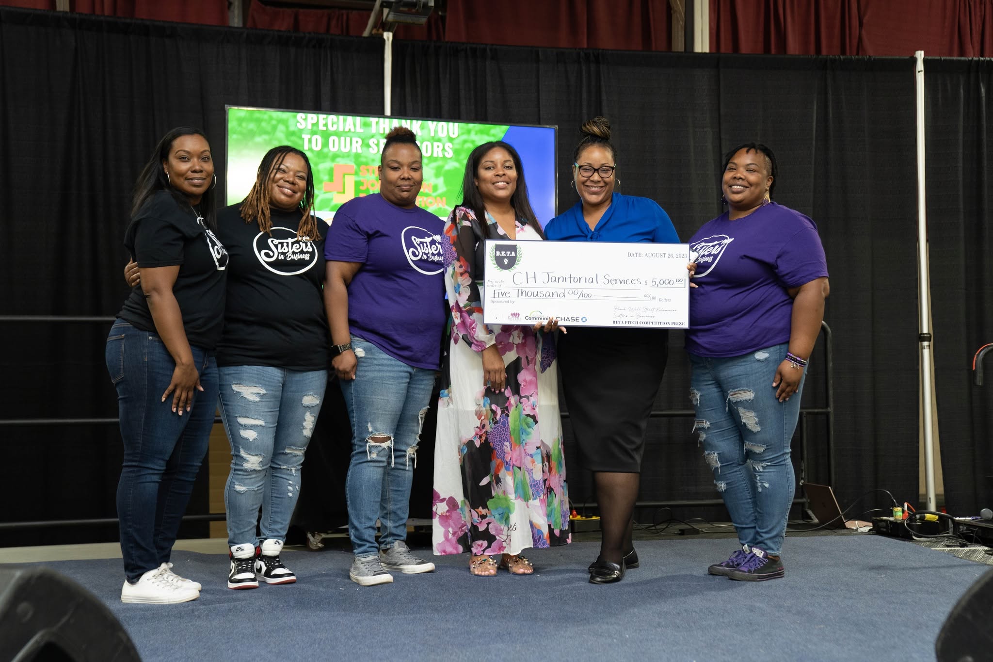 Six women standing on stage, holding a large check, smiling, with a black curtain background and a green screen behind them.