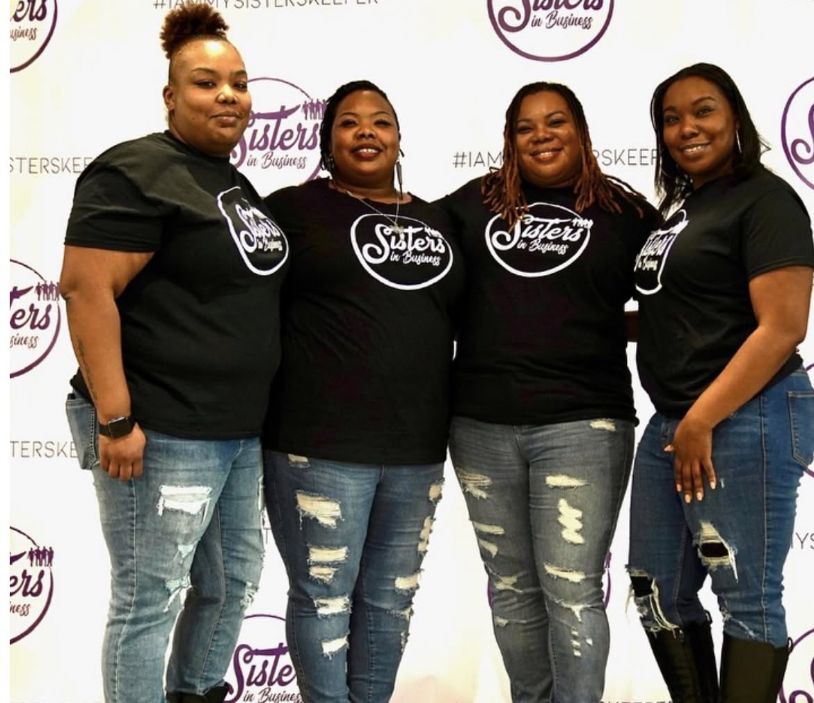 Four women stand together in front of a backdrop with logos, wearing matching black shirts and ripped jeans.