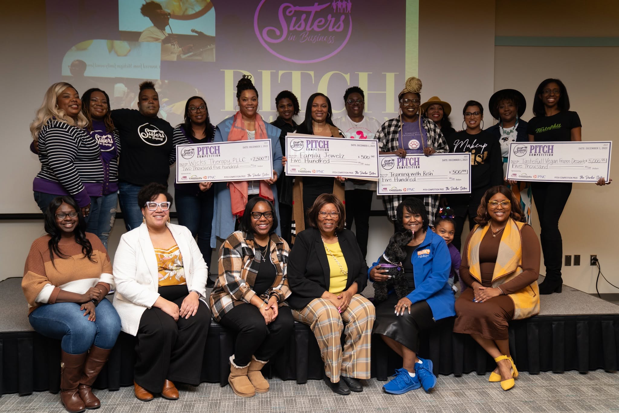 Group of women and girls on stage holding large checks, some sitting and some standing, with a screen and banner in background.