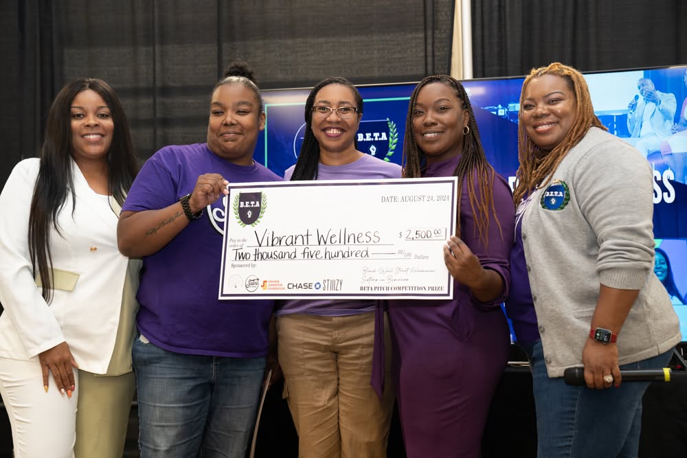 Group of five women holding a large check, smiling, indoors with a dark curtain background.