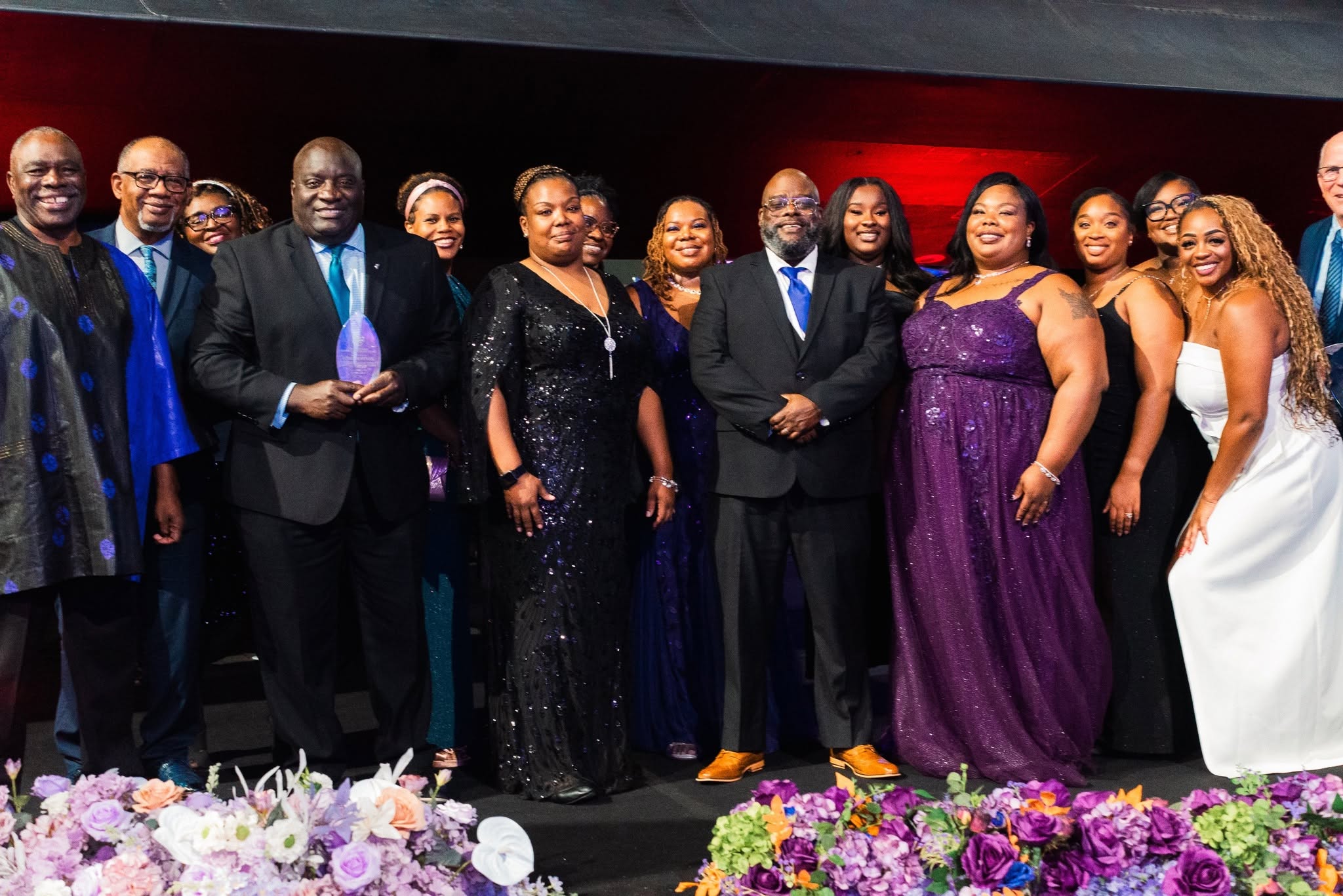Group of people dressed in formal attire standing on stage with flowers in front, smiling for a photo.
