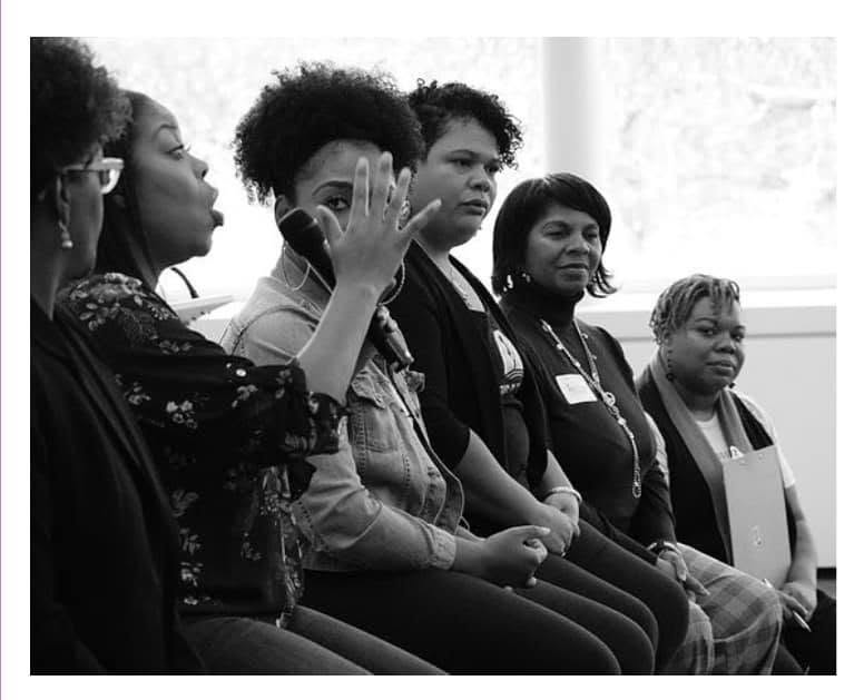 Five women sitting in a row, one raising her hand, in a room with a window behind them.