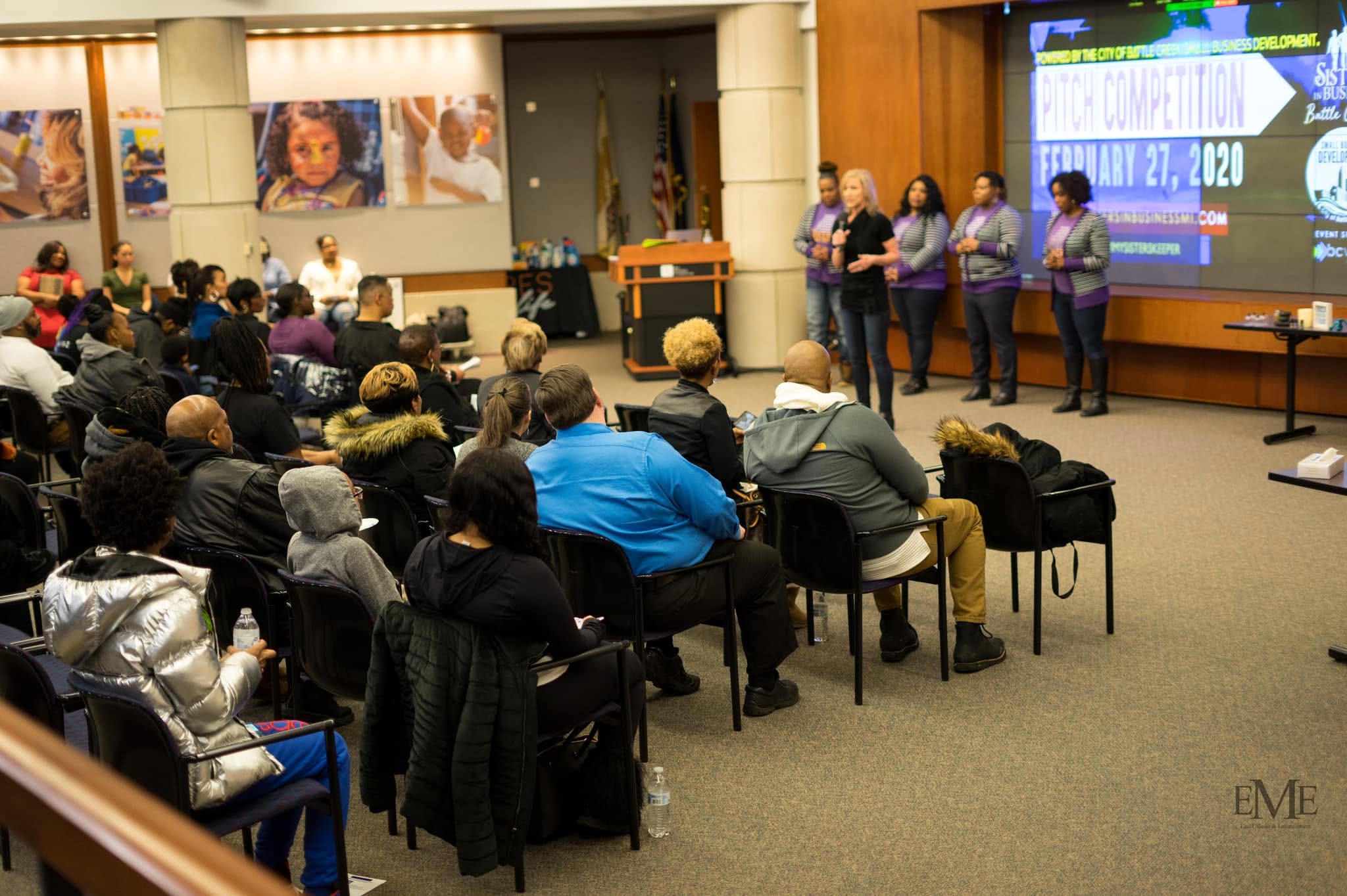 Group of people seated in a conference room watching a presentation on stage with five people speaking, large screen displaying text, and posters on wall.