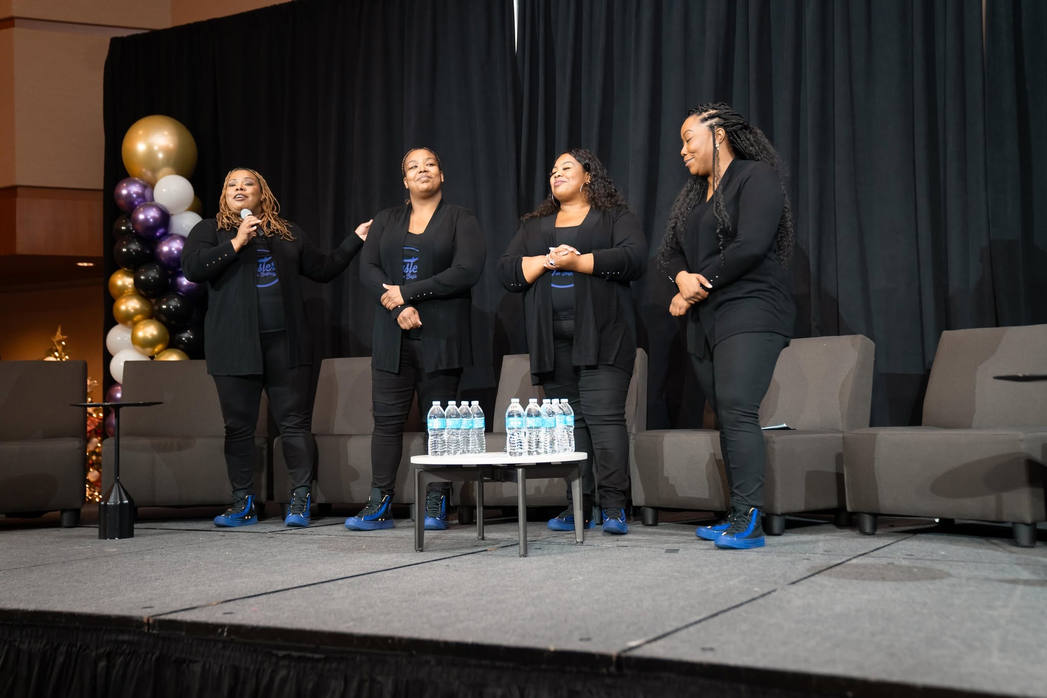 Four women stand on stage with a table of water bottles, black curtains, and balloons in the background.