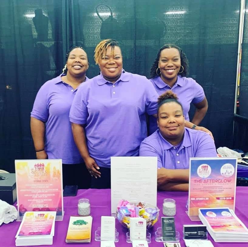 Four women in purple shirts standing behind a table with informational materials, smiling, in front of a dark curtain.