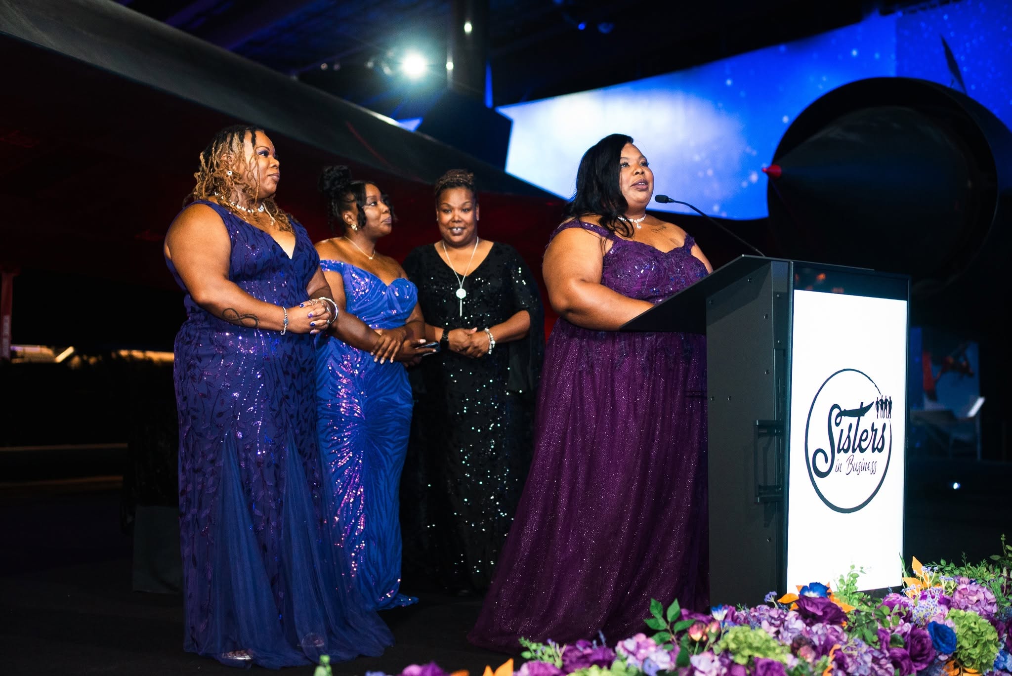 Four women in formal dresses stand on stage behind a podium with a floral arrangement, speaking at an event.