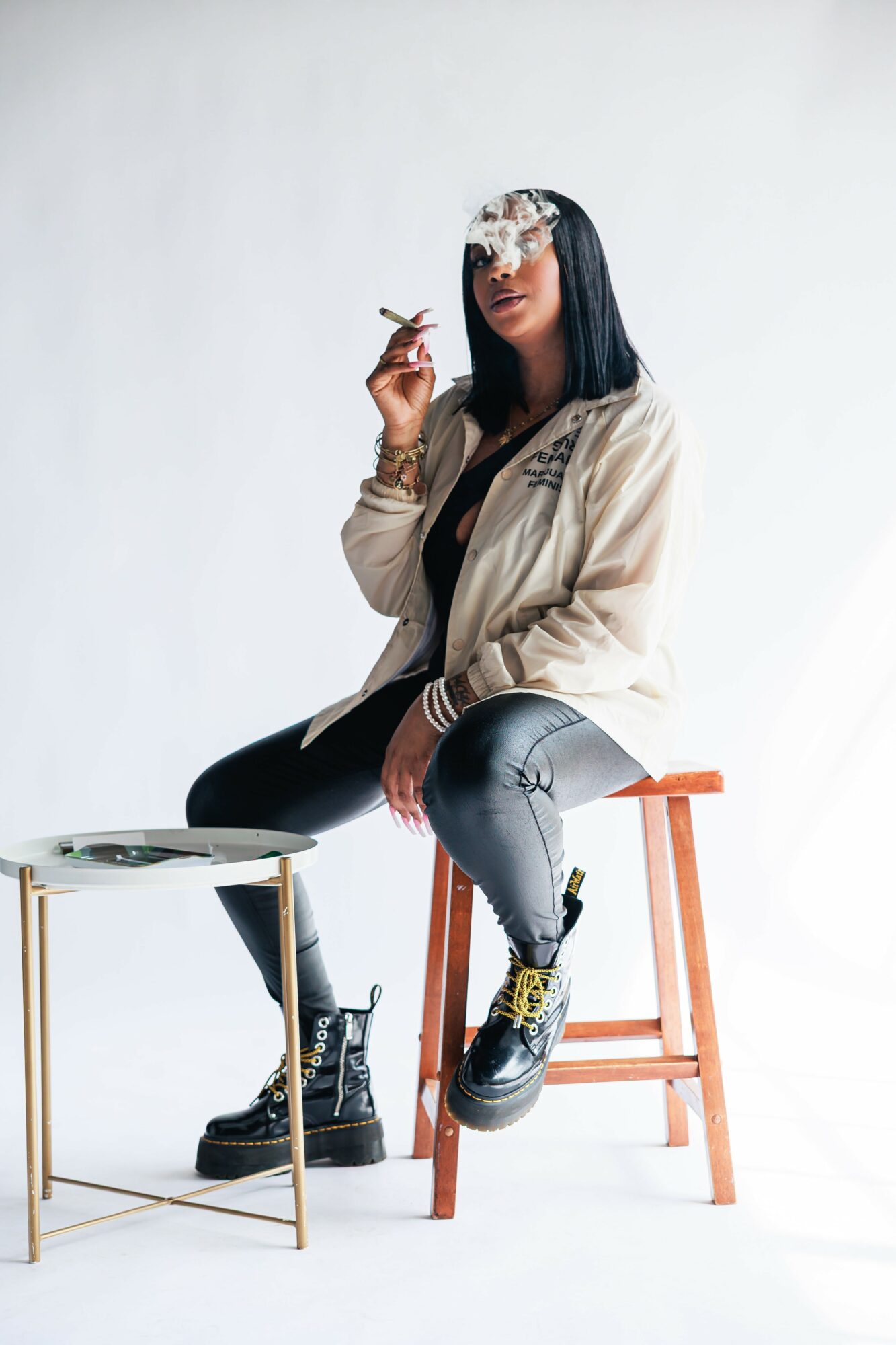 Woman sitting on a wooden stool, smoking, with a small table nearby, against a plain white background.