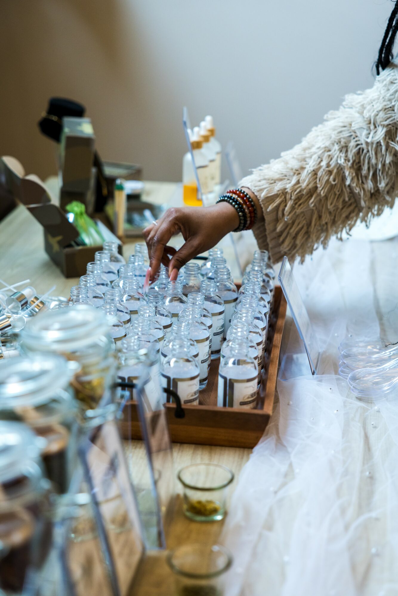 Person's hand reaching for small glass bottles on a table with various items, including jars and containers, in the background.