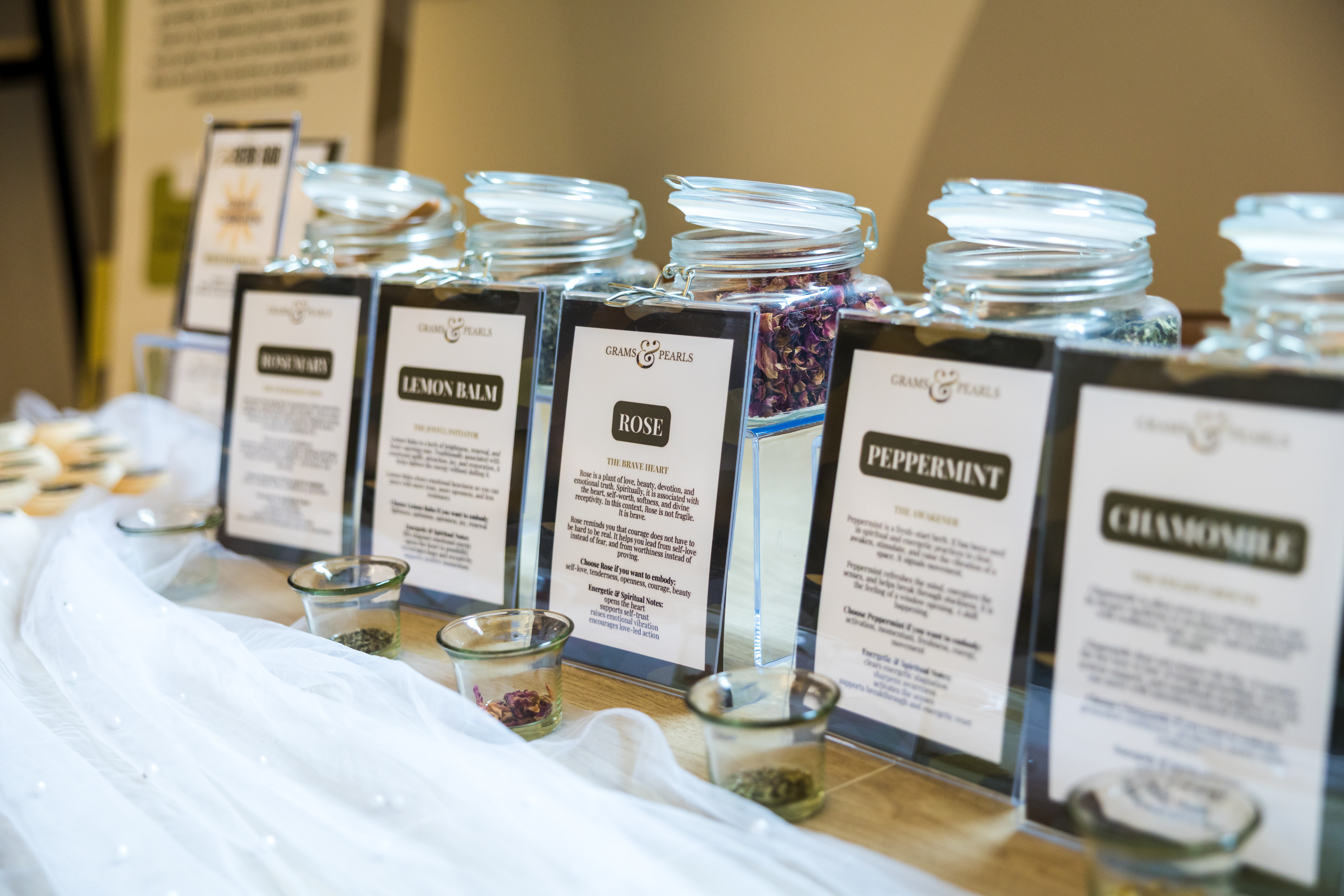 Display of glass jars with labels and small scoops on a table, with informational signs in front, in a well-lit setting.