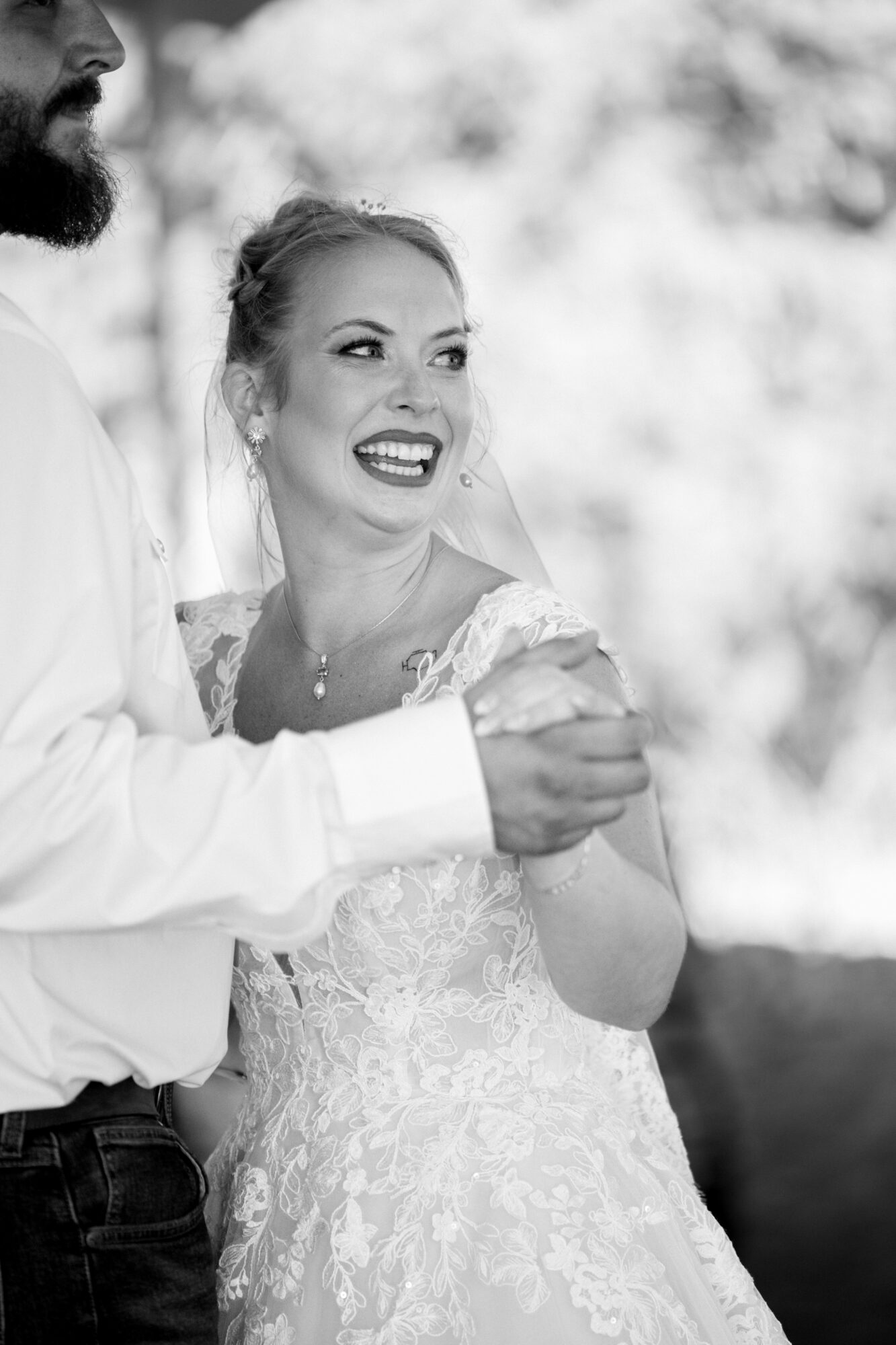 Smiling woman in wedding dress with a person, outdoors, blurred background, black and white, joyful expression.