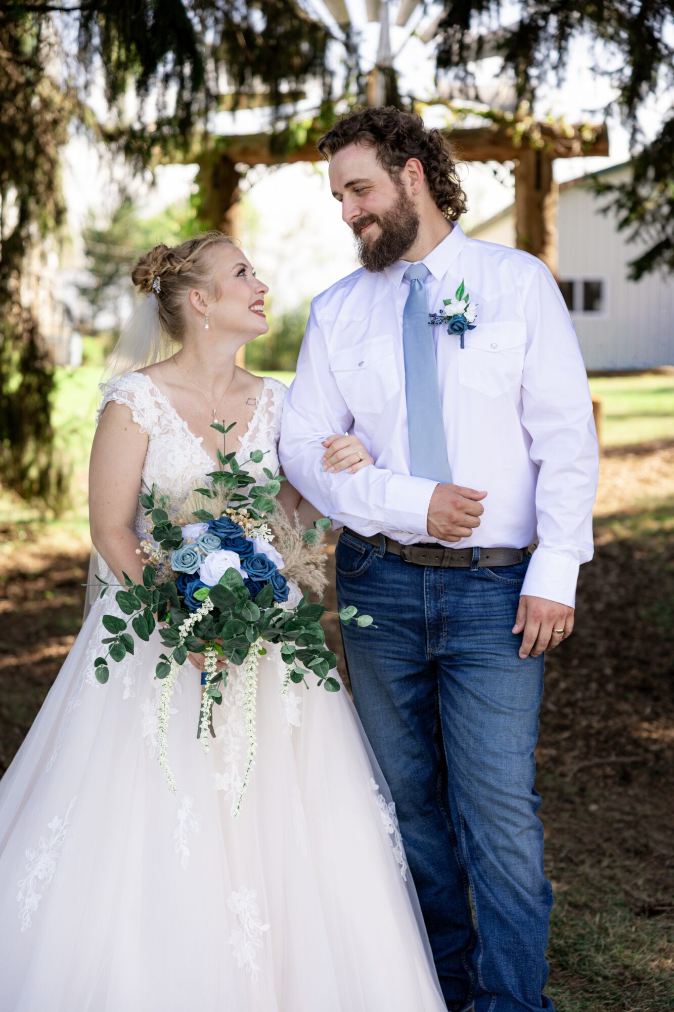 A bride and groom stand outdoors, smiling at each other, with trees and a wooden arch in the background.