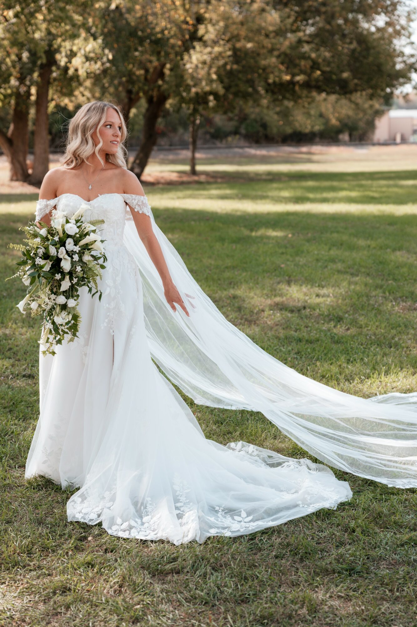 Bride in a white wedding dress holding a large bouquet outdoors on grass with trees in background.