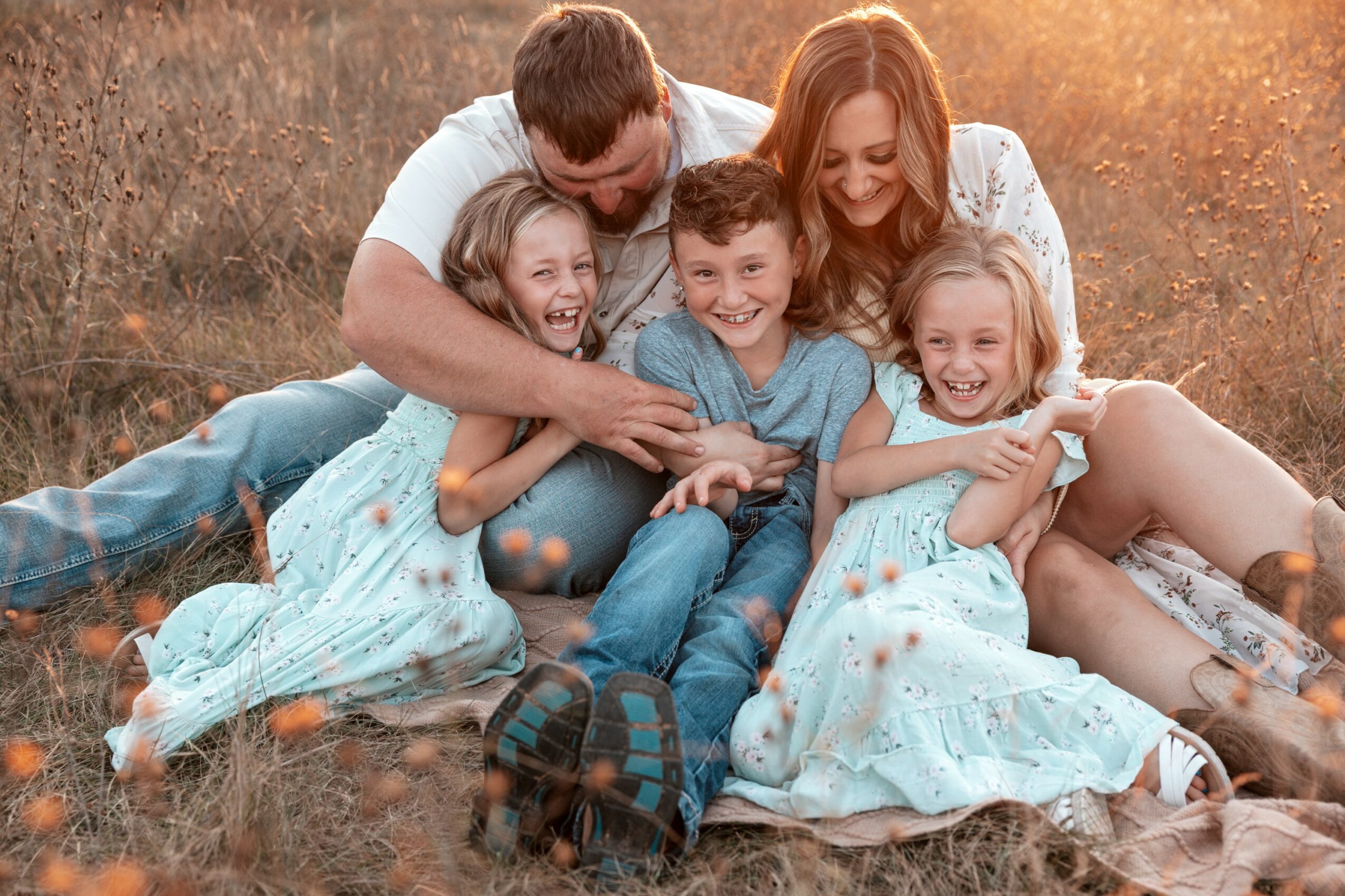 Family of five sitting on grass outdoors, smiling and hugging each other, warm sunlight in background.