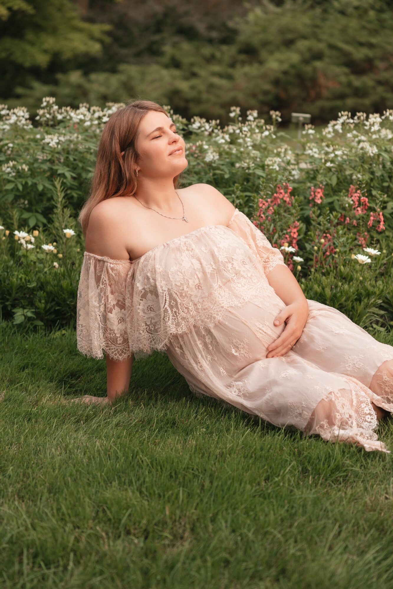 Pregnant woman in a light dress sitting on grass in a garden with flowers, eyes closed, smiling, sunlight on her face.