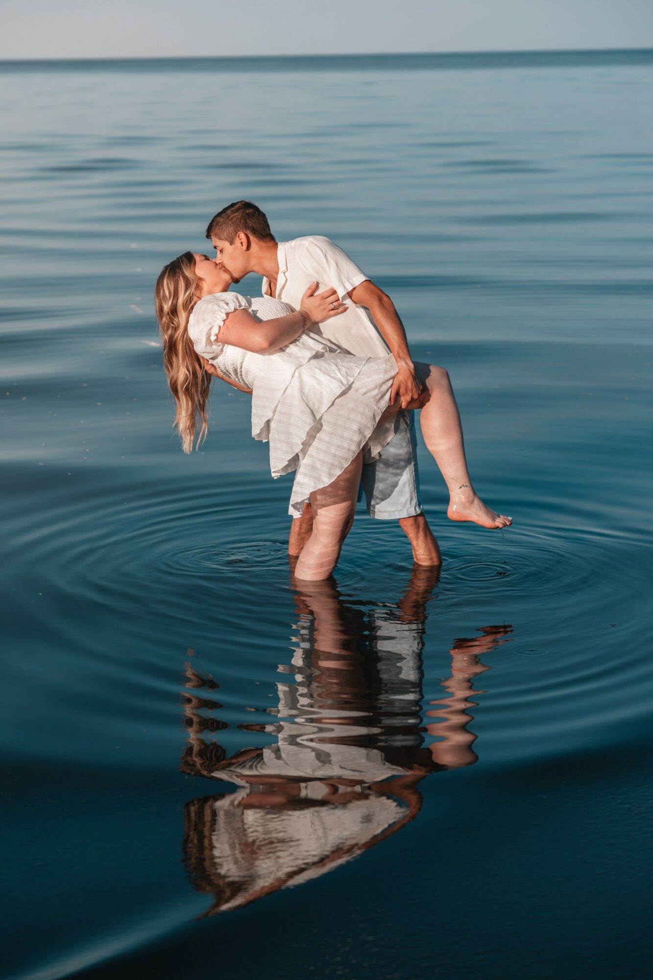 A couple stands in water, embracing and kissing, with a calm body of water and horizon in the background.