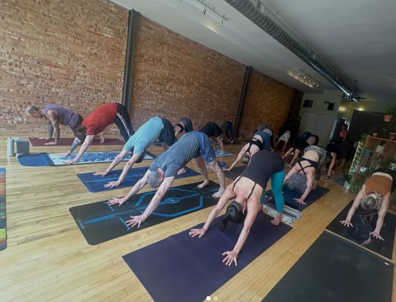 People practicing yoga on mats in a studio with brick walls and wooden floors, in a downward dog pose.