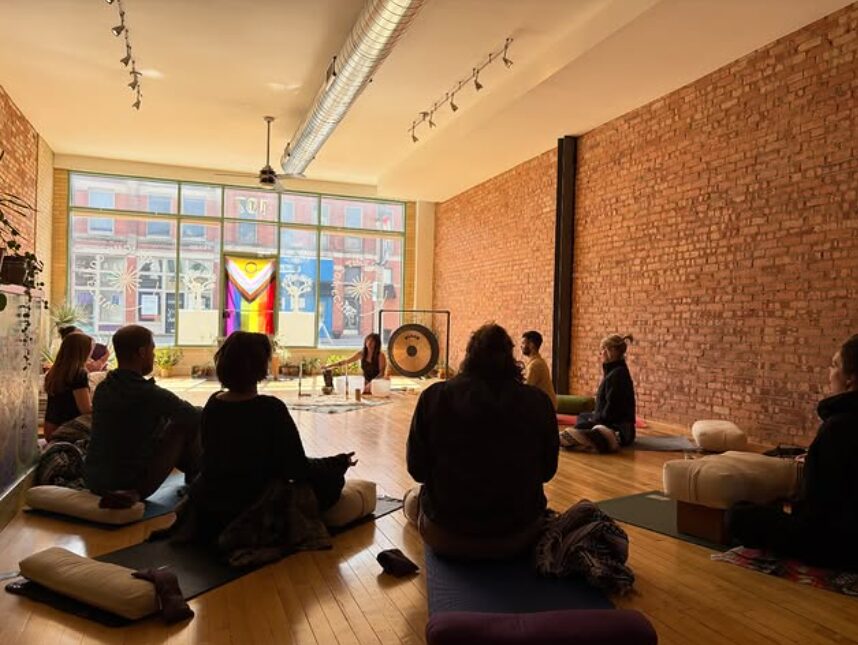 People sit on cushions in a spacious room with brick walls, facing a gong and a colorful window, during a meditation session.