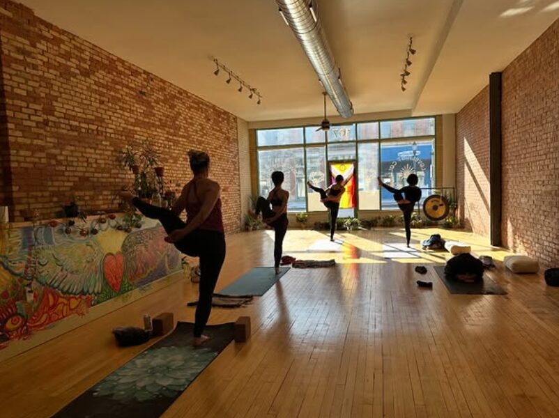 Group of people practicing yoga in a studio with brick walls and large windows, sunlight streaming in.