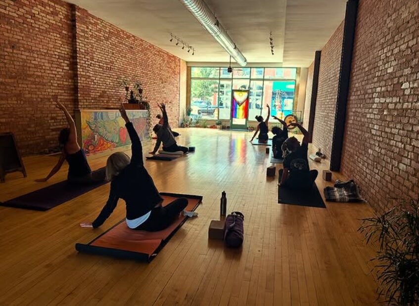People practicing yoga in a spacious studio with brick walls and large windows, some seated and some stretching on mats.