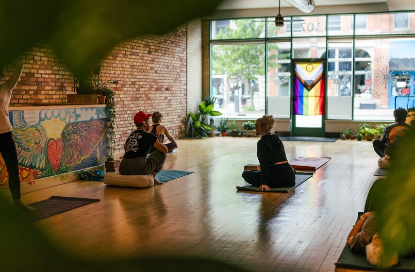 People practicing yoga in a bright, spacious room with large windows, brick wall, and colorful artwork, during daytime.