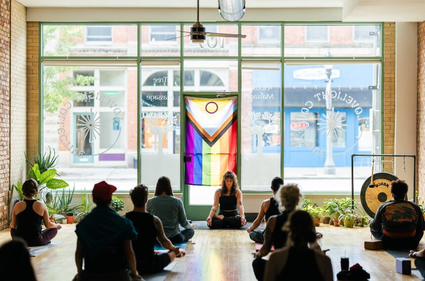 Group of people sitting on the floor in a room with large windows and colorful artwork, practicing meditation or yoga.