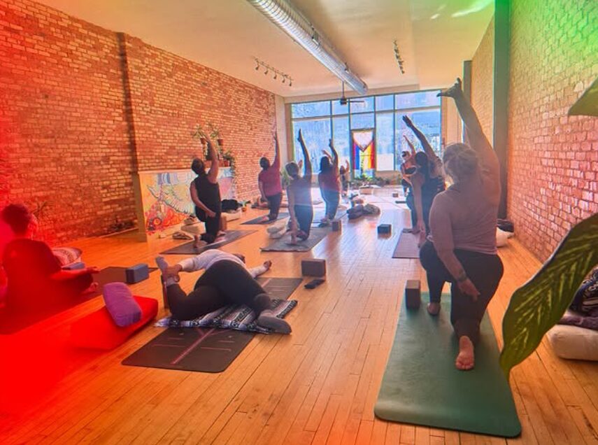 People participating in a yoga or stretching class in a bright, spacious room with brick walls and large windows.