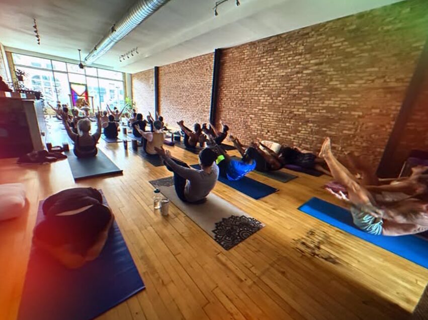 People practicing yoga in a spacious studio with large windows and brick walls, sitting on mats with arms raised.