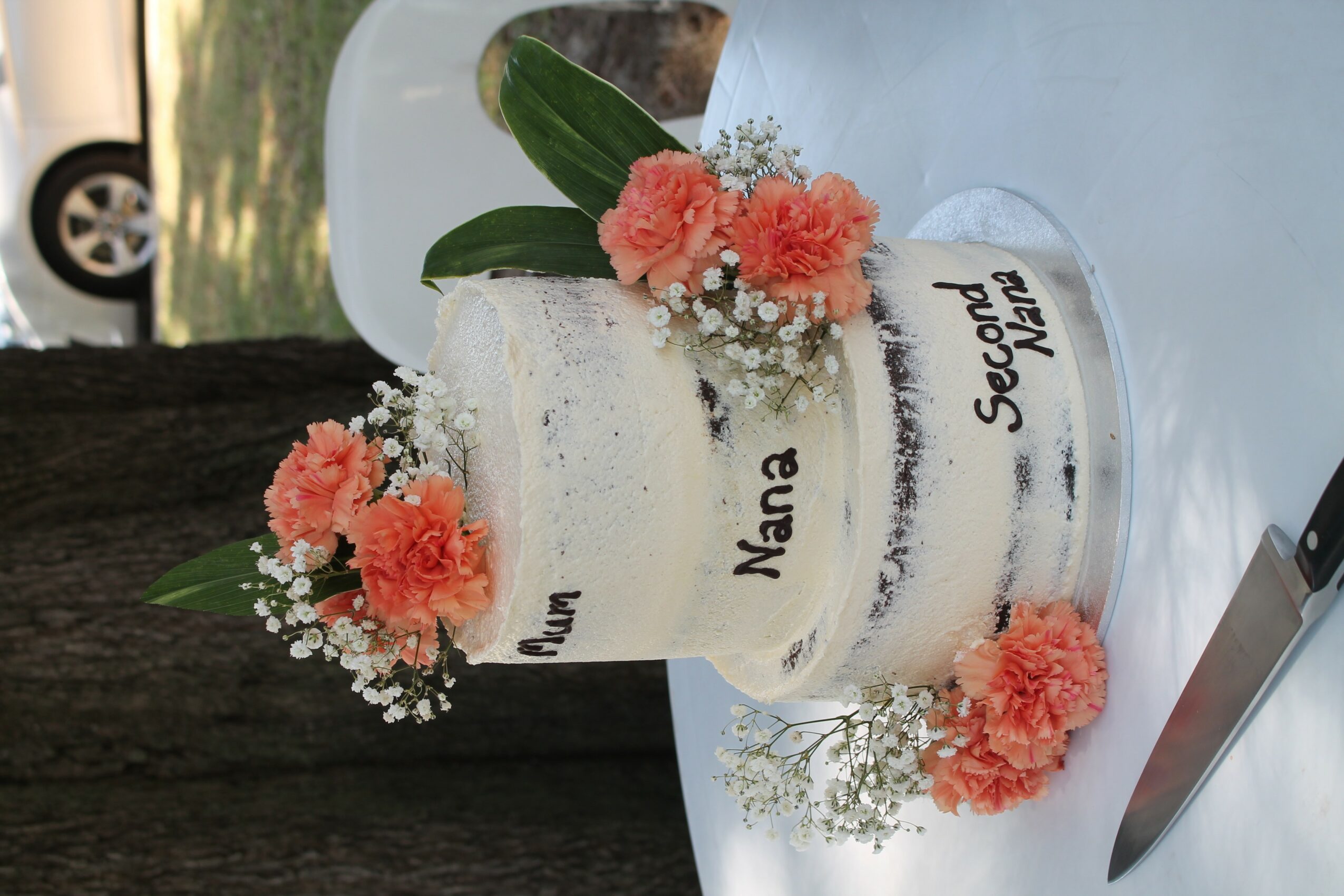 Two-tier white cake with pink flowers and green leaves, on a glass stand, with a knife nearby.