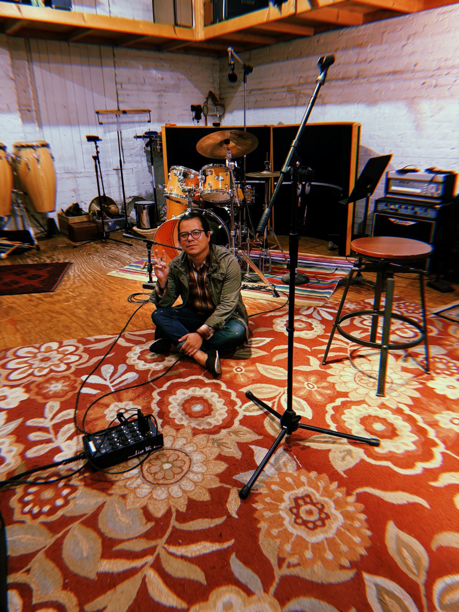 Person sitting cross-legged on a patterned carpet in a music studio with drums, microphones, and chairs around.