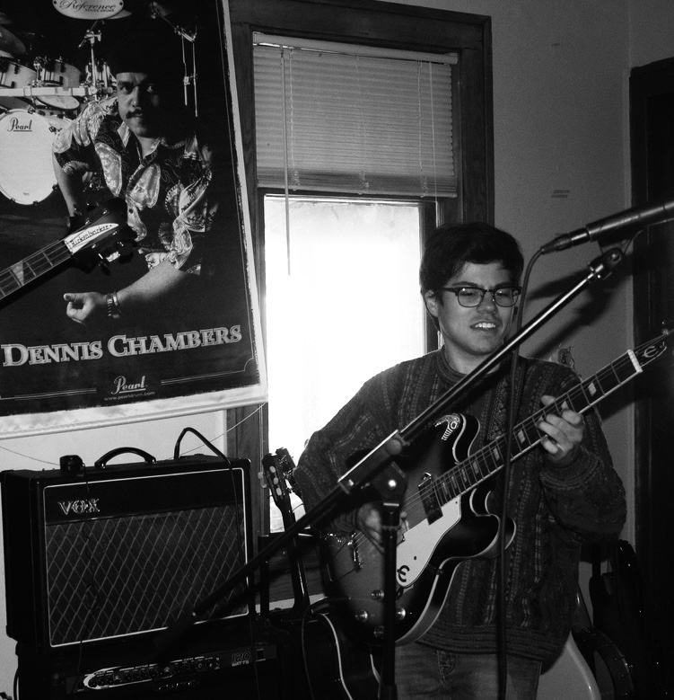 Young man with glasses playing an electric guitar in a room with a window and a poster of Dennis Chambers, with music equipment around.