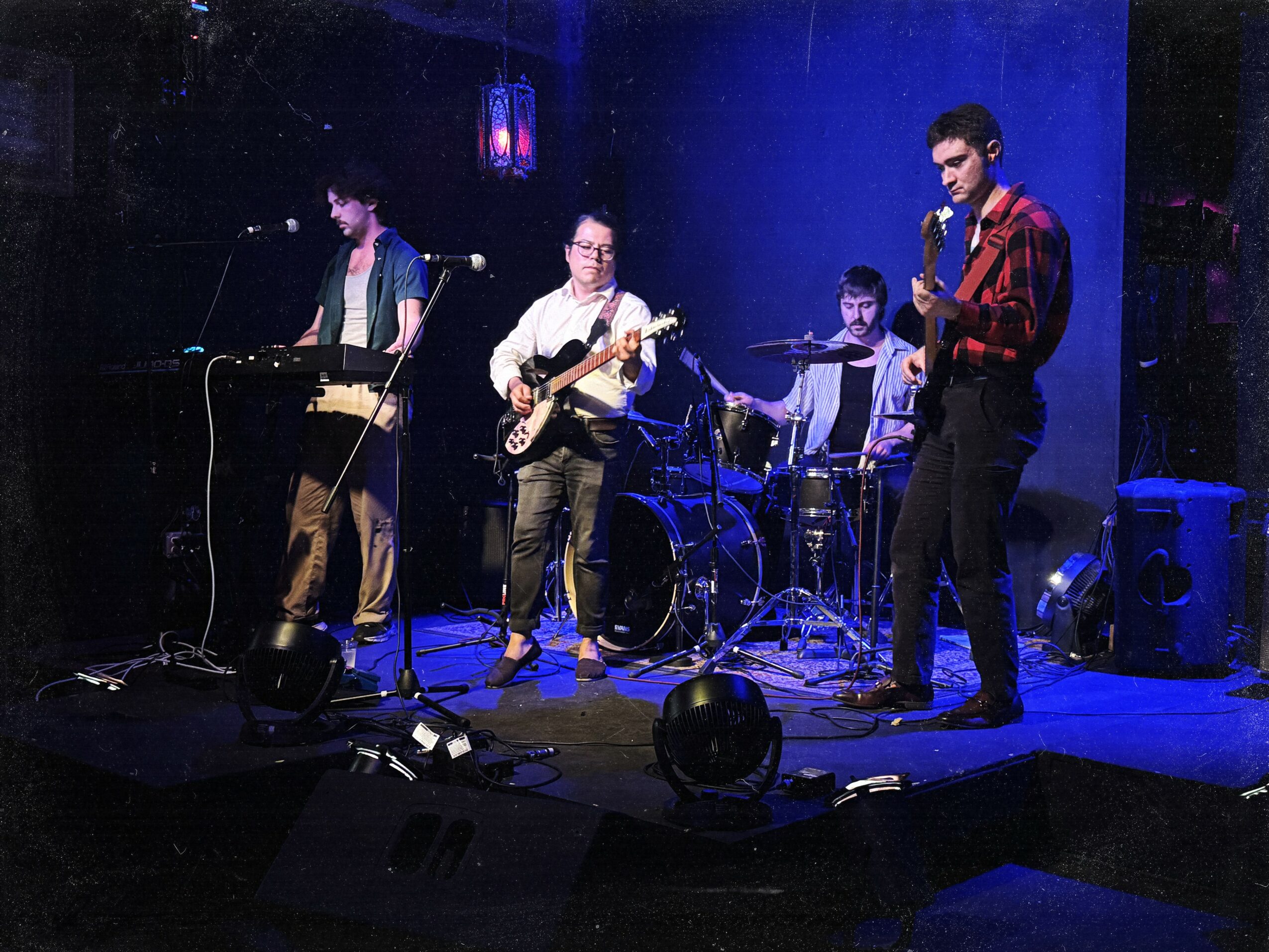 Four musicians perform on stage with instruments, illuminated by blue and purple lighting, in a dark venue.