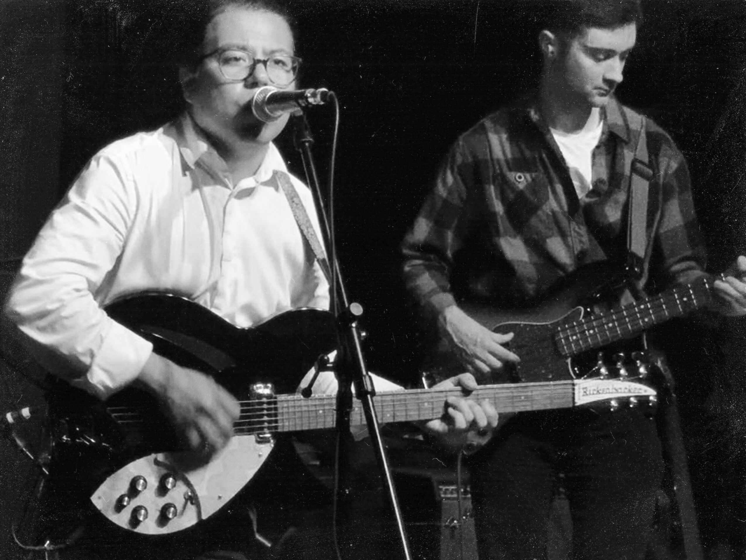 Two people playing electric guitars, one singing into a microphone, in a black and white setting.