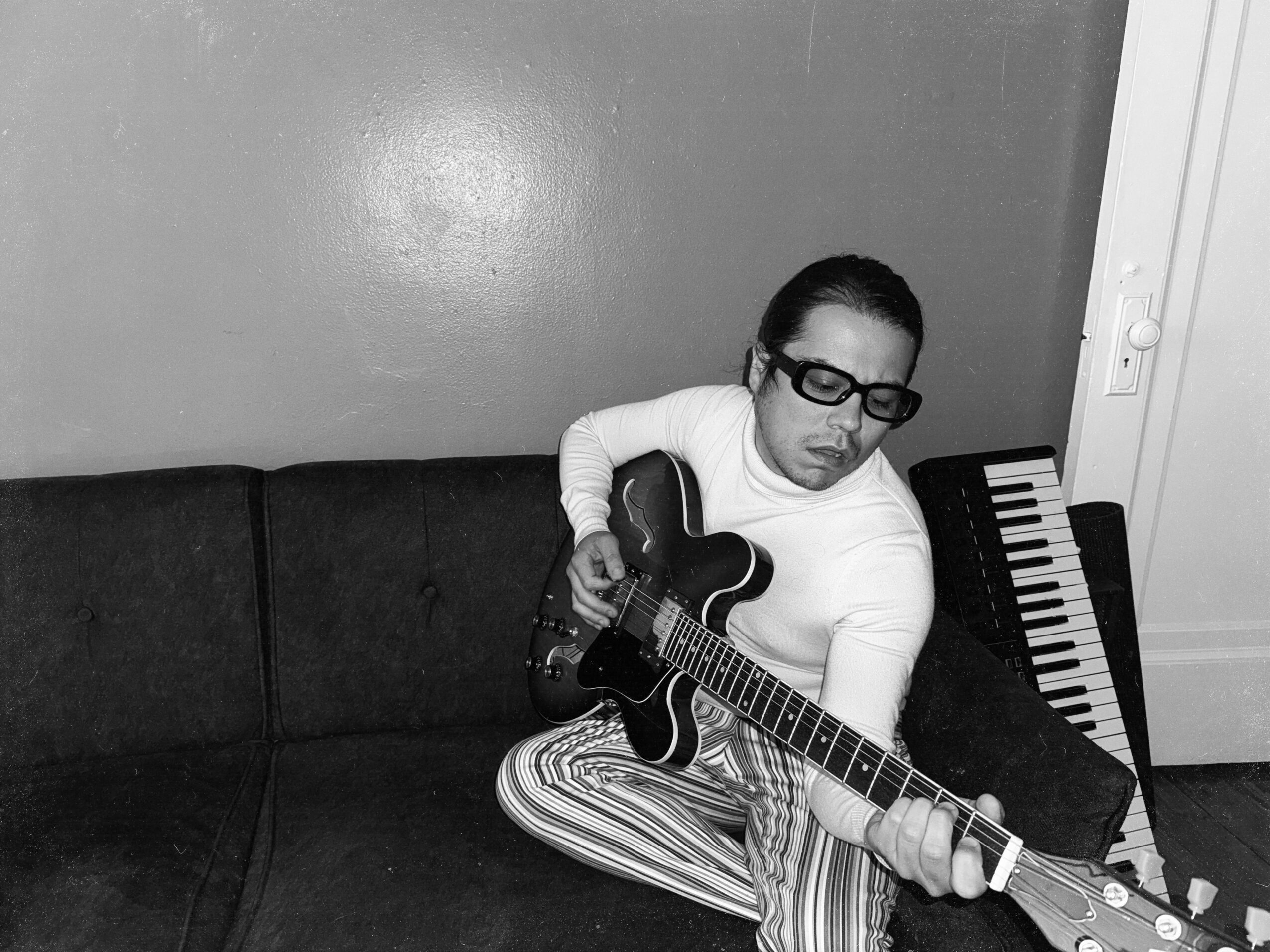 Person sitting on a couch playing an electric guitar, with a keyboard nearby, in a room with plain wall.