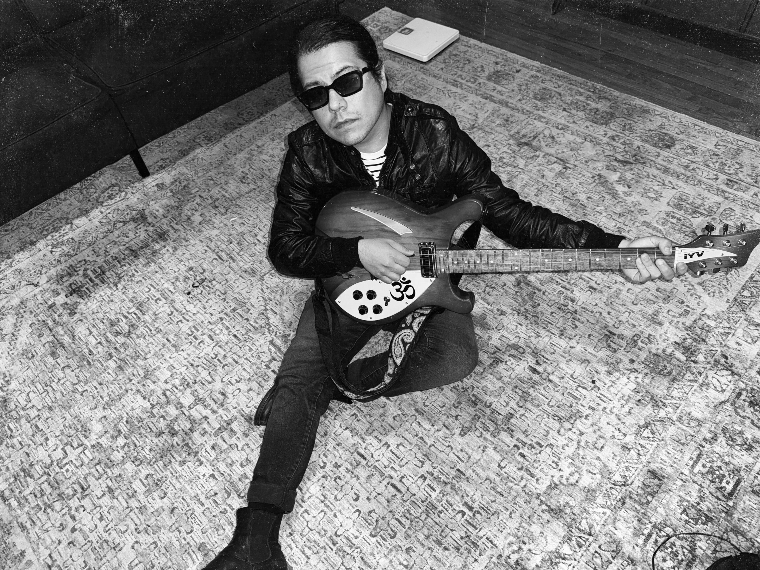 Person sitting on a rug holding a guitar, wearing sunglasses and a leather jacket, with a book nearby.