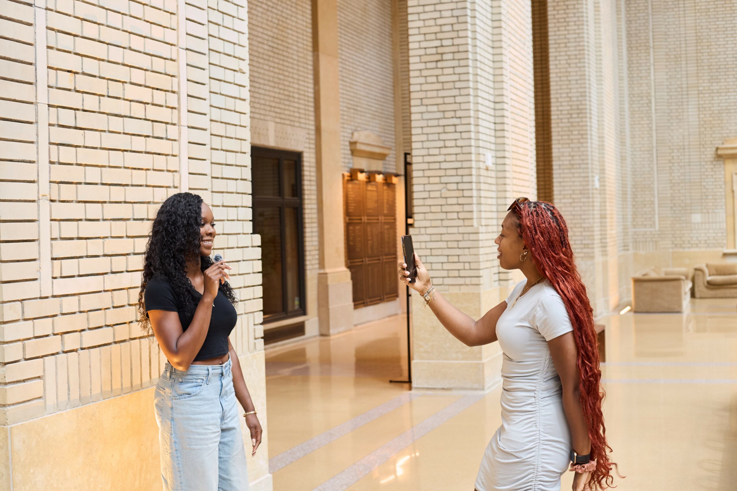 Two women stand in a spacious, well-lit indoor area with brick walls, one taking a photo of the other.