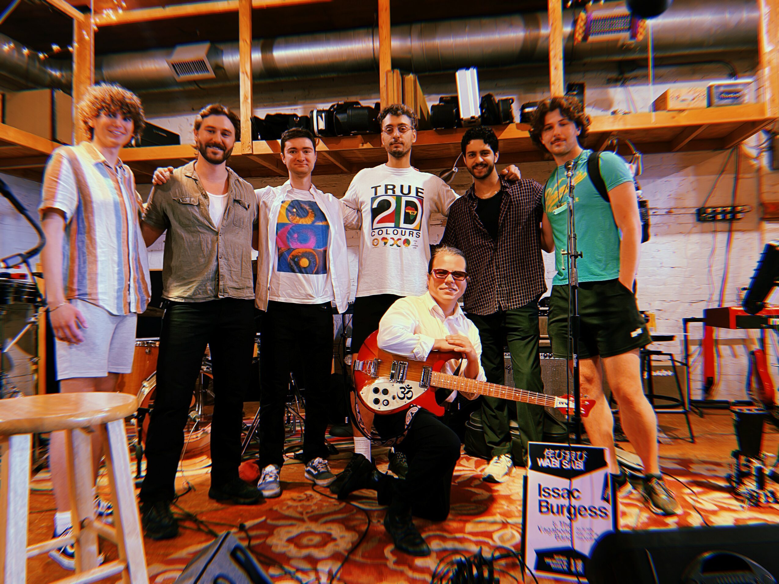 Group of seven people, one holding a guitar, in a room with wooden shelves and equipment, smiling at the camera.