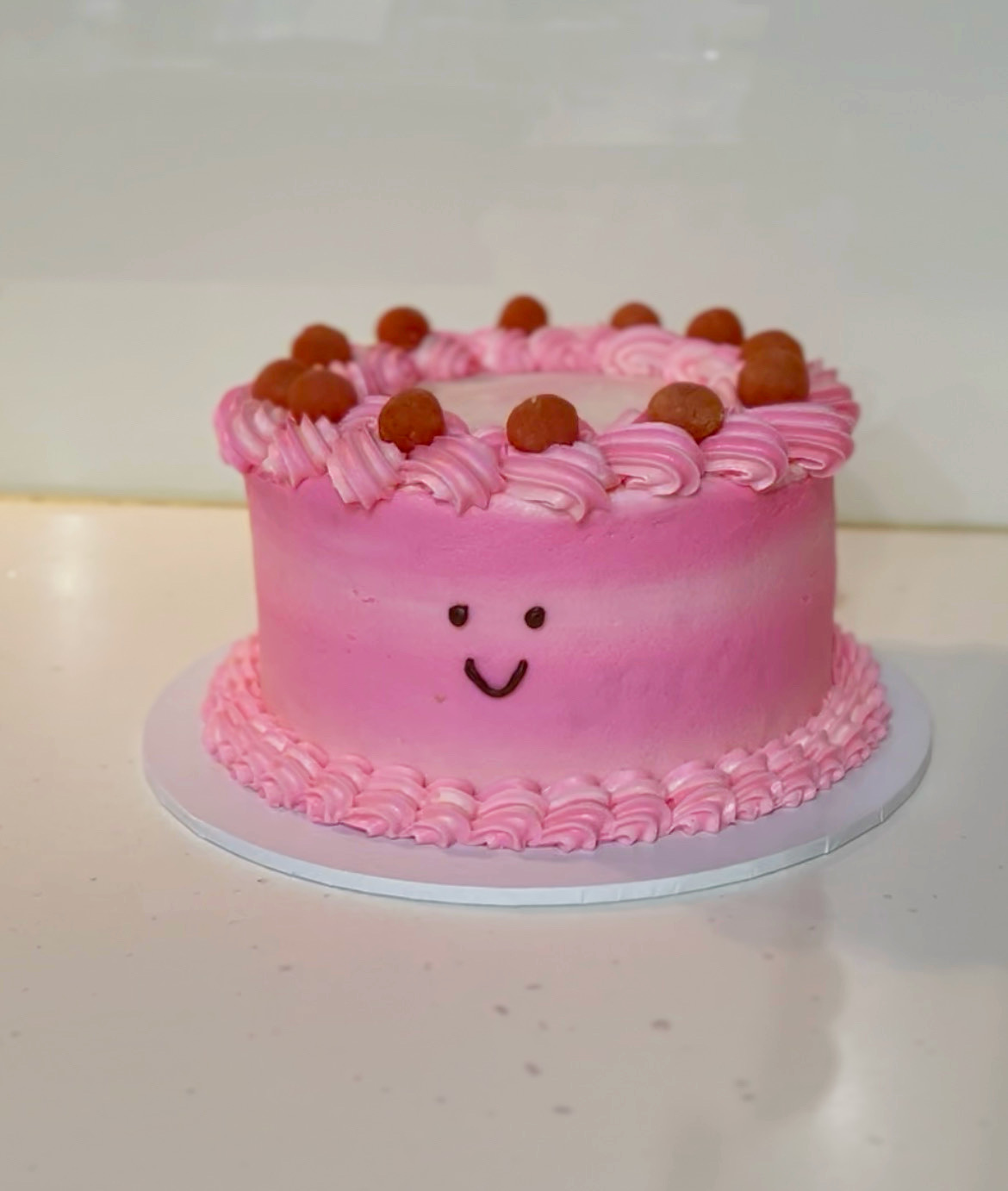 Pink cake with a smiling face, decorated with pink frosting and small round toppings, on a white surface.