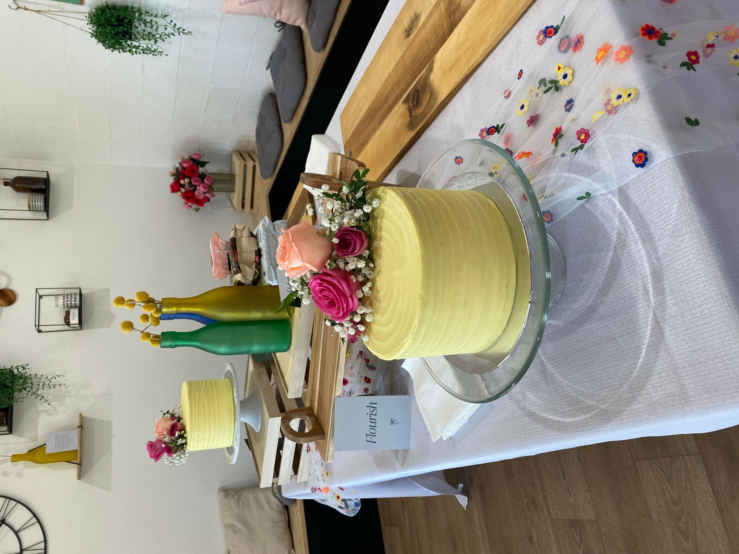 A yellow cake with pink and purple flowers on top, decorated with a small card, on a table with a white tablecloth.