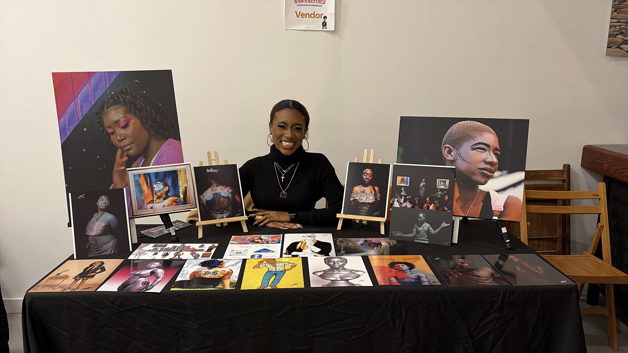Person sitting at a table with artwork and photographs displayed, smiling, in an indoor setting.