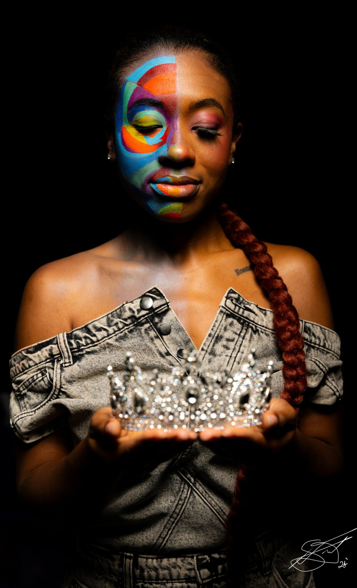 Young woman with colorful face paint, holding a crystal crown, wearing an off-shoulder patterned top, against a black background.