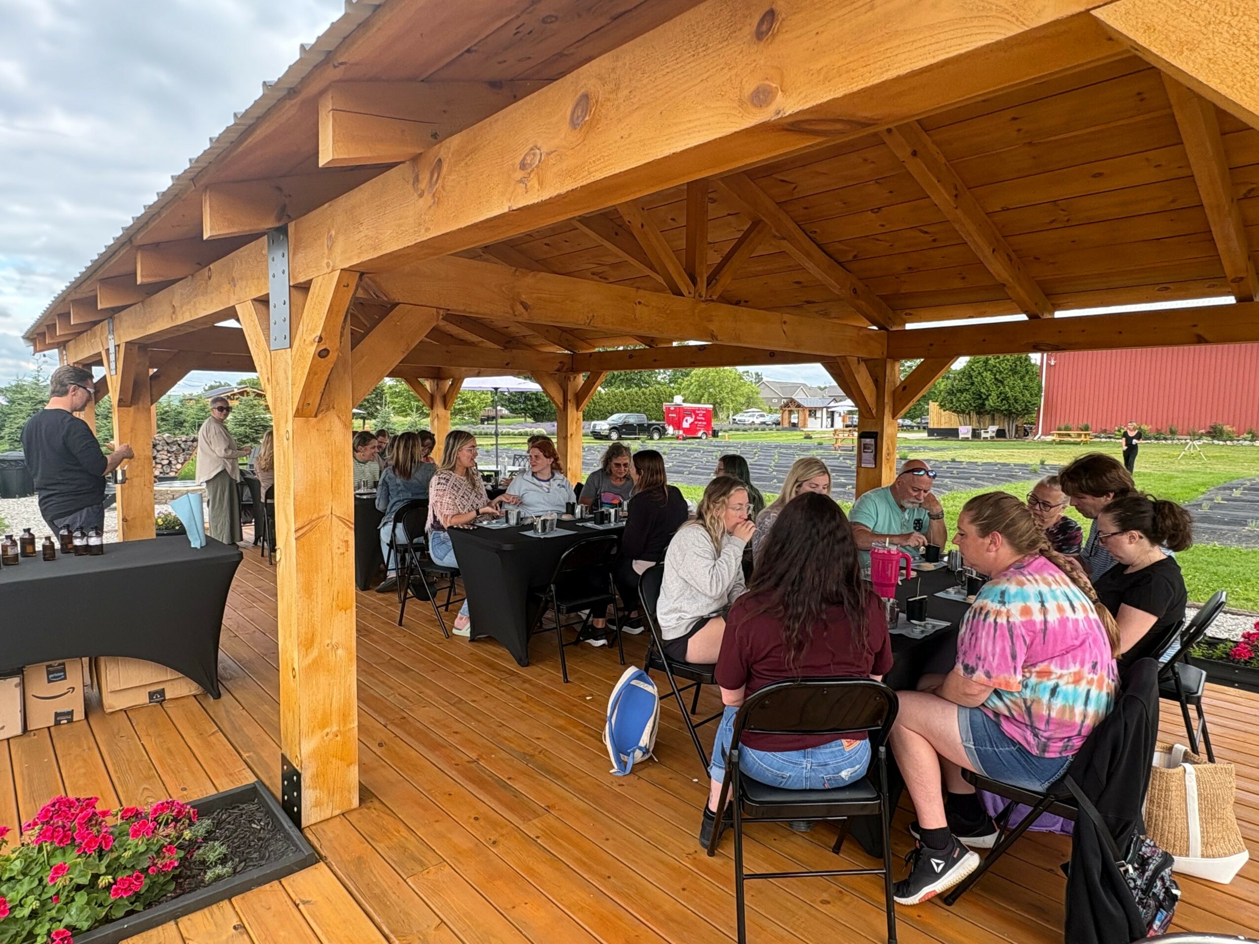 People sitting at tables under a wooden pavilion outdoors, with a grassy field and trees in the background.