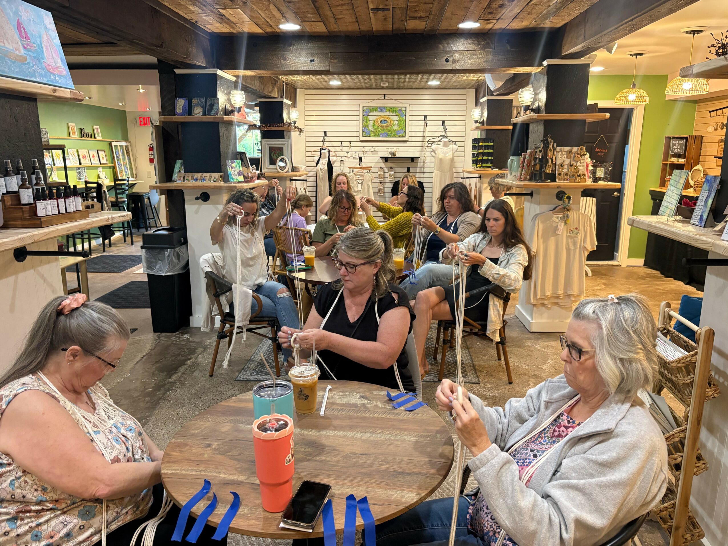 Group of women sitting at tables in a cozy cafe, engaging in conversation and activities, with shelves and decor in the background.
