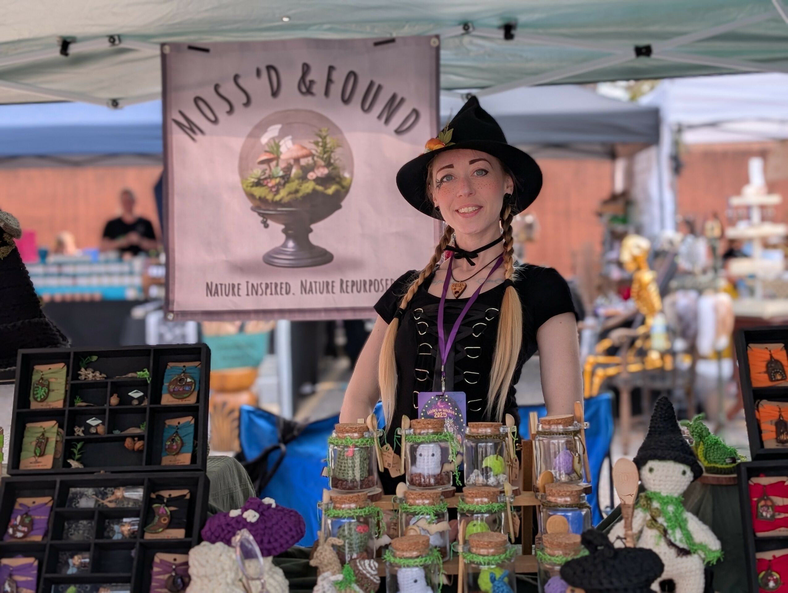 Woman in witch costume at outdoor market stall with jars and crafts, under a tent with a 'Moss'd & Found' sign.