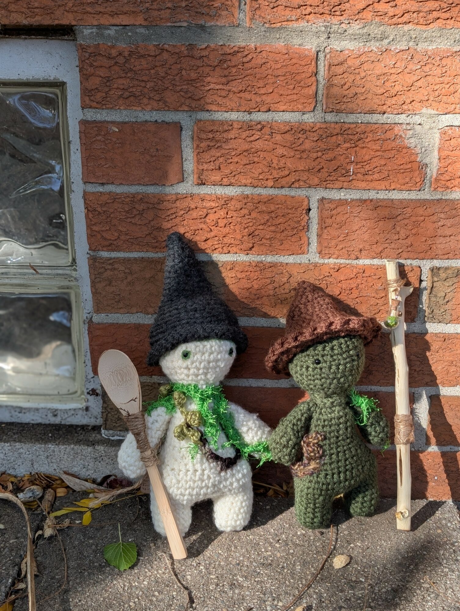 Two knitted dolls dressed as witches with hats and wands, standing on a sidewalk against a brick wall.