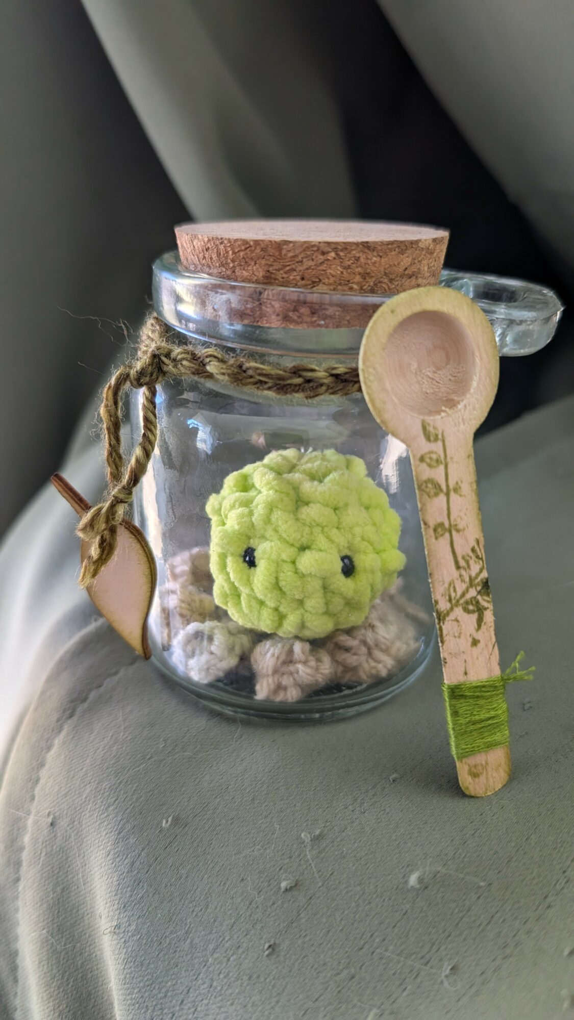 Small glass jar with a cork lid, decorated with a twine bow, contains a green plush toy, a wooden spoon, and small objects inside.