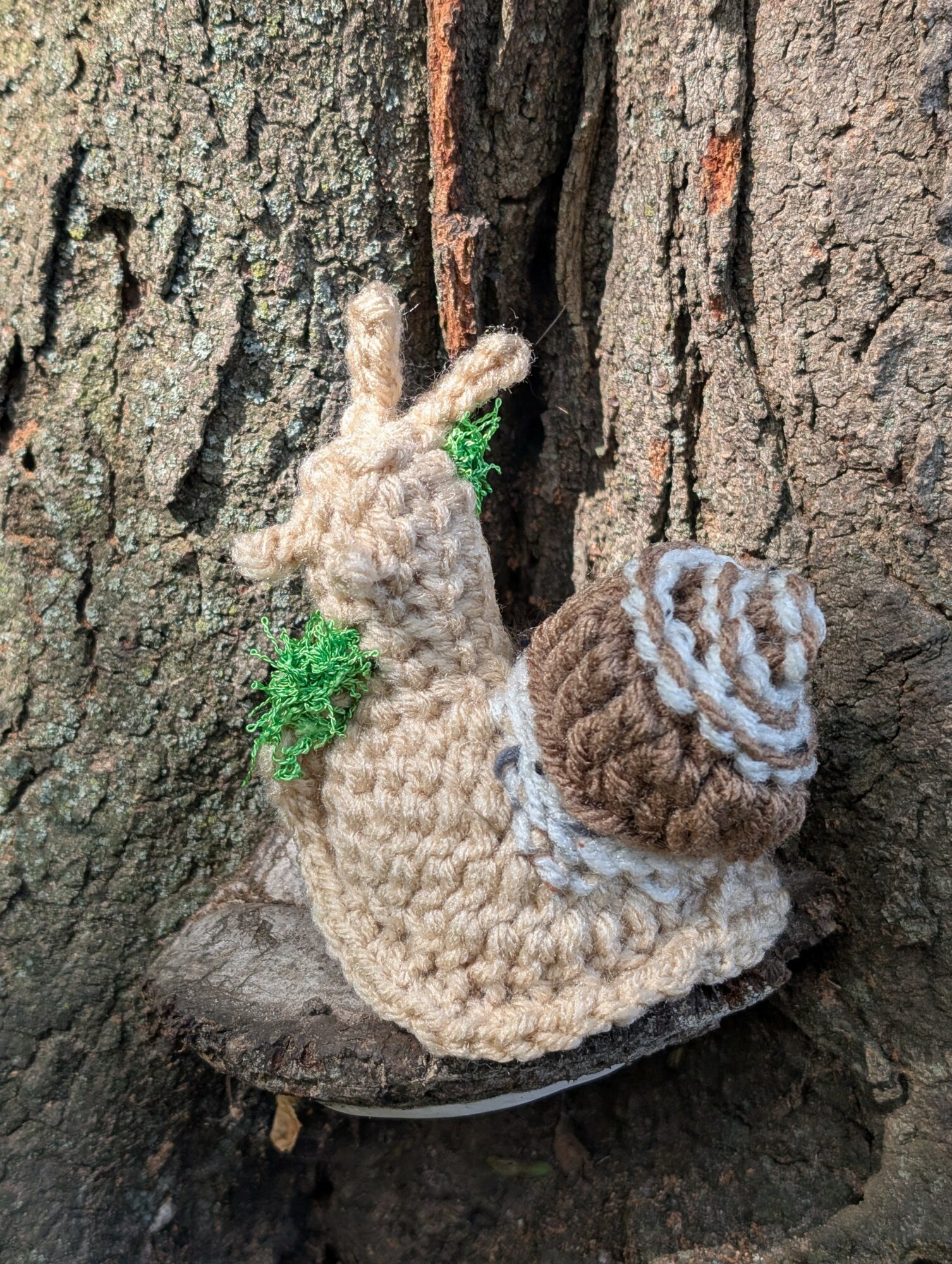 Knitted snail with a brown and white shell, green foliage, on a tree trunk, close-up view.