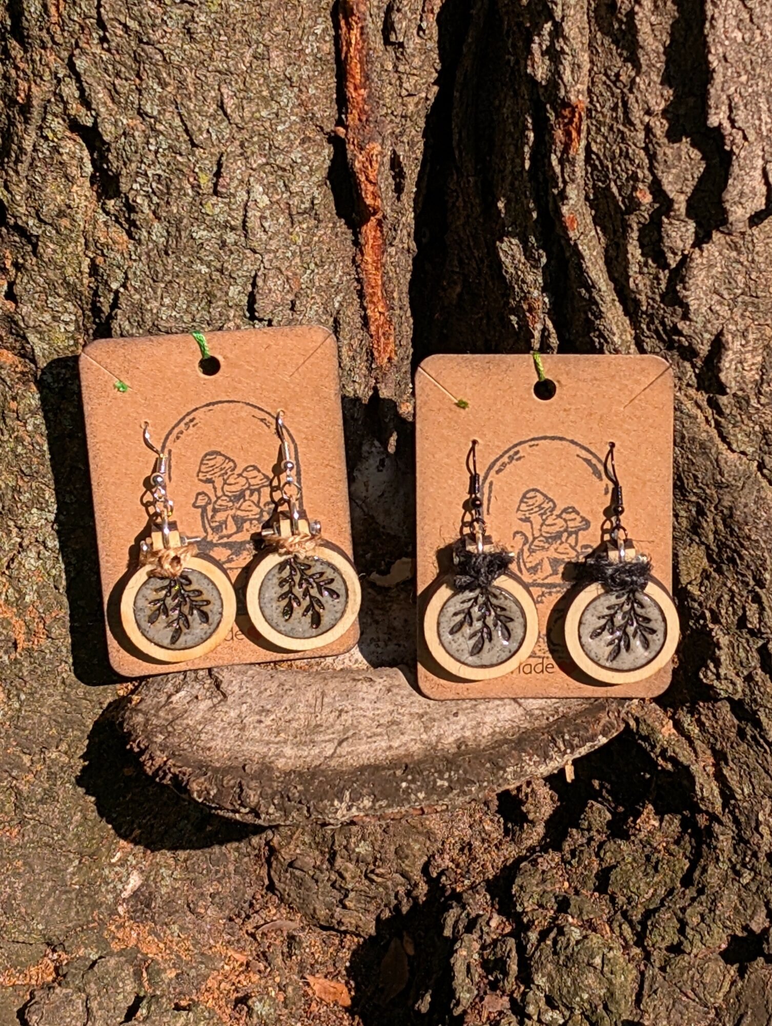 Two pairs of earrings with tree designs hang on brown cards, displayed against a tree bark background.