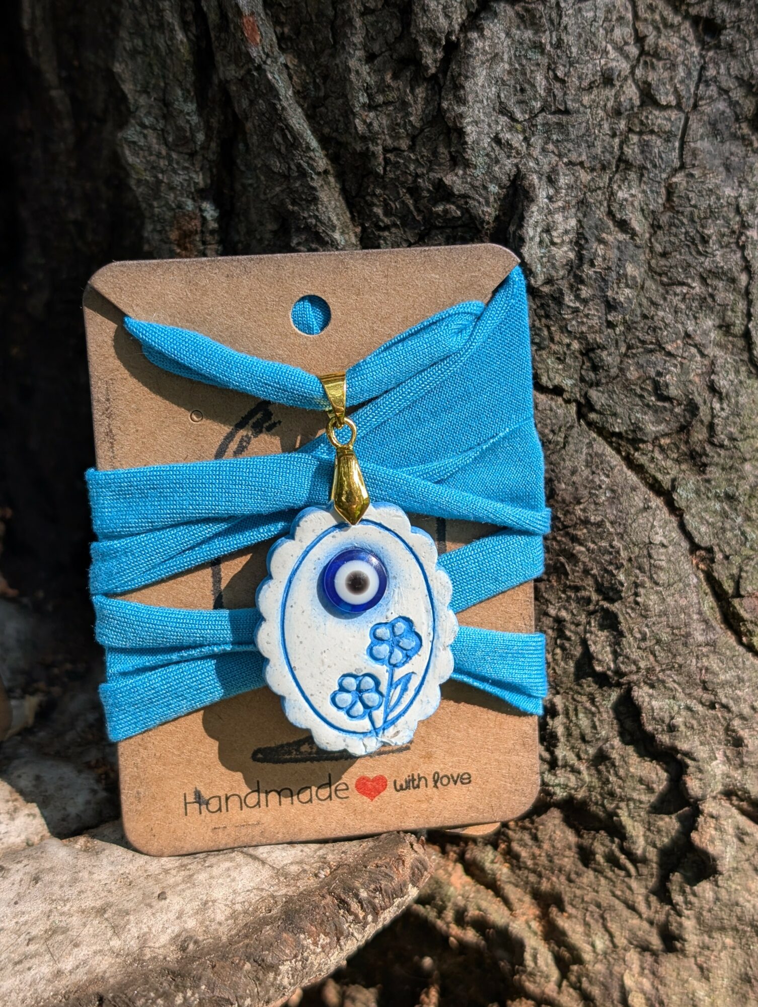 Blue fabric hair tie with a white and blue Nazar amulet charm attached, displayed on a cardboard backing against a tree bark background.