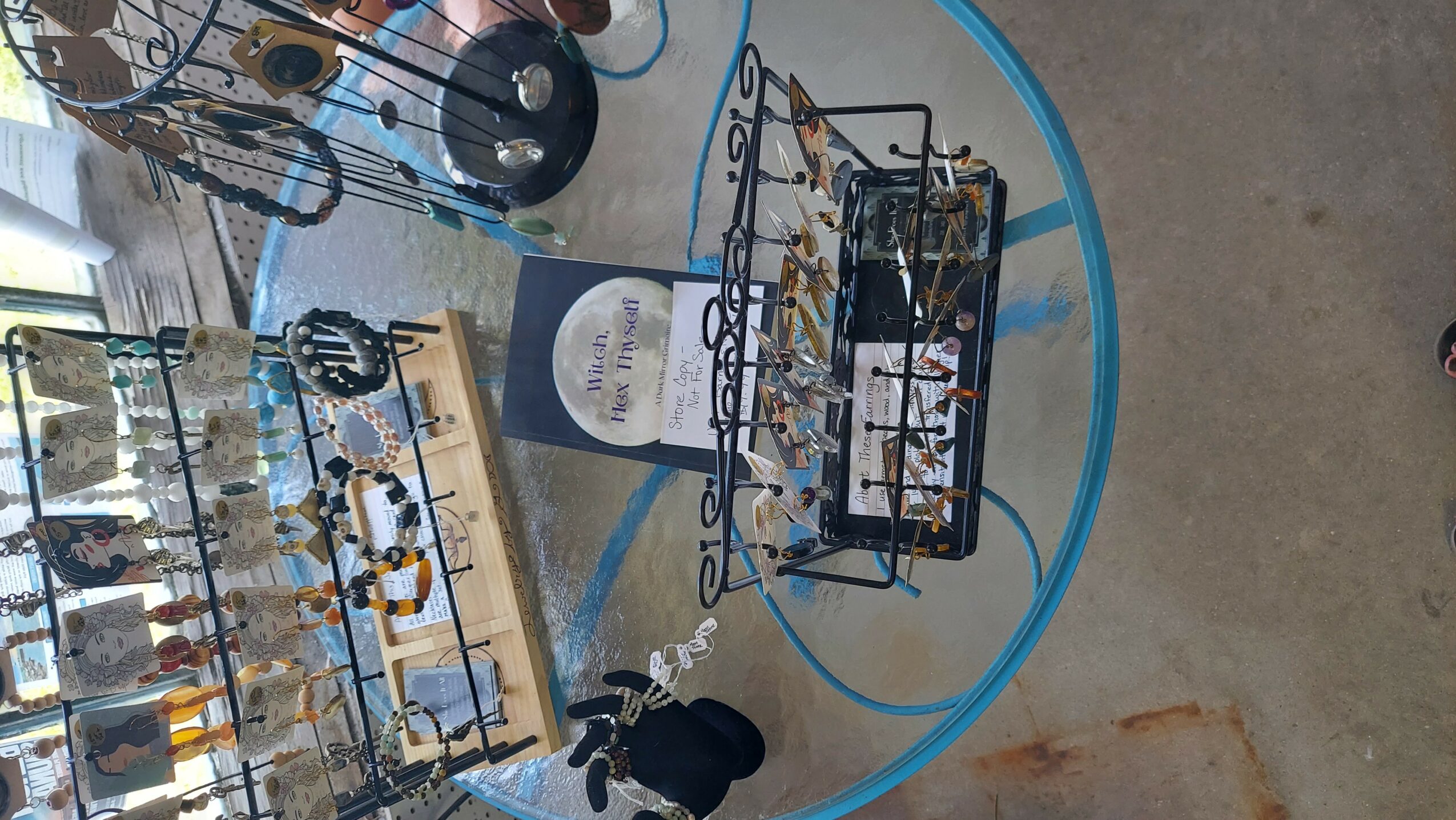 Display of jewelry and accessories on tables at an indoor market or store, with a blue circular table and concrete floor.