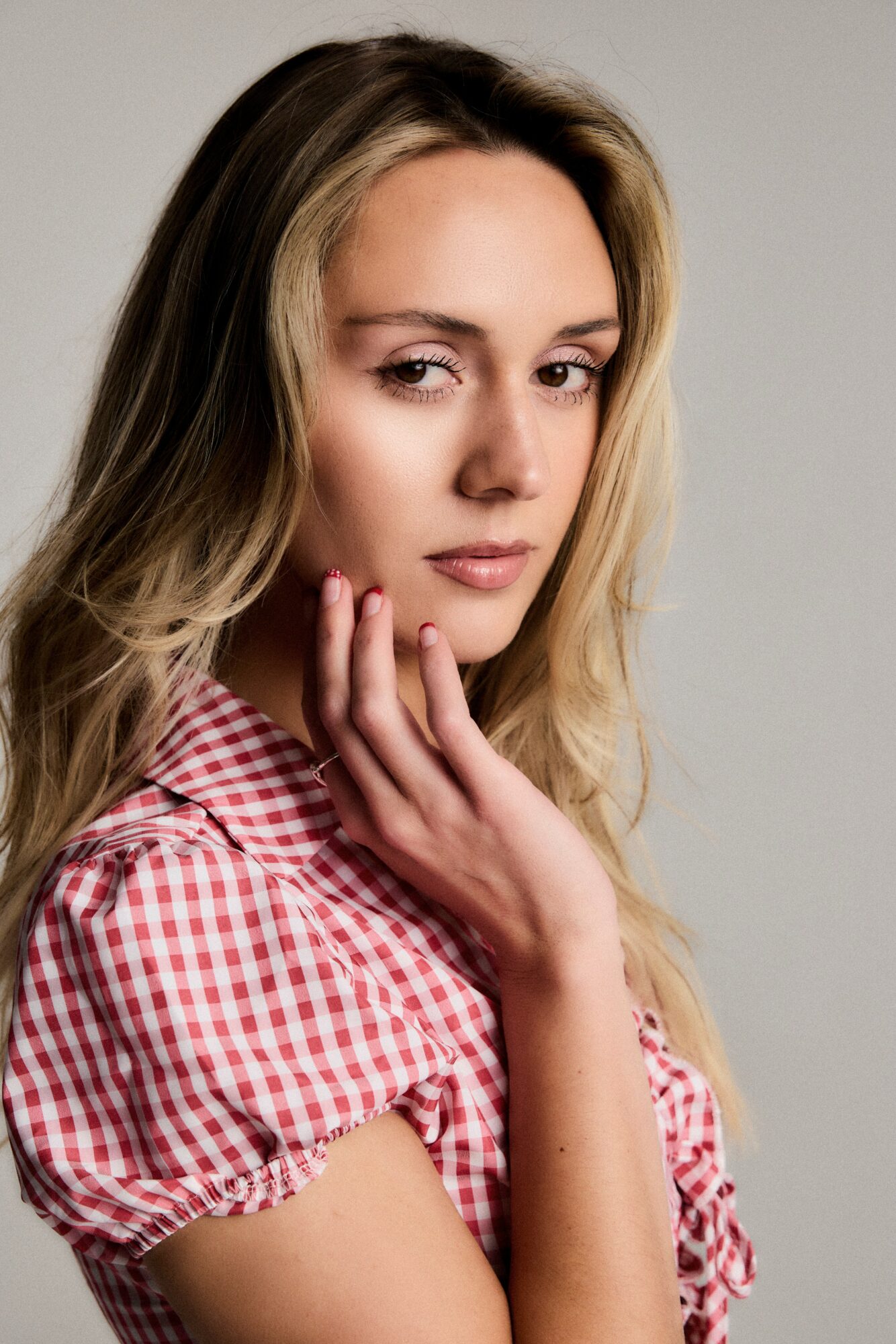 Young woman with long blonde hair, wearing a red and white checkered shirt, touching her face gently, looking at the camera.